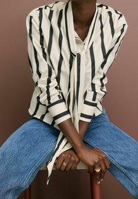 Hvid bluse med sorte vertikale striber, der har en bundet krave og lange ærmer, parret med blå denimjeans. Glat stofstruktur.