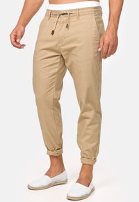 Pantaloni casual beige con vita regolabile, vestibilità affusolata, orli arrotolati e due tasche frontali. Abbinati a scarpe bianche slip-on.