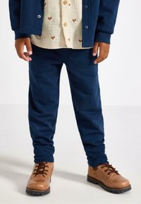 Kind in marineblauwe broek, beige shirt met vosgezichten, marineblauwe jas en bruine veterschoenen staat op een lichte vloer.