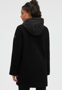 Black fleece coat with a contrasting black padded hood, long sleeves, and a textured, soft exterior, featuring a side logo detail.