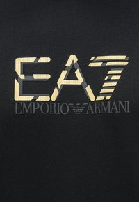 EA7 logó arany texturált betűkkel, Emporio Armani felirattal és sas emblémával fekete szövet háttéren.