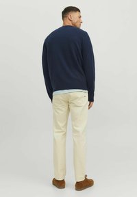 Pulover marin cu guler rotund, asortat cu pantaloni bej deschis. Modelul stă cu spatele la cameră, purtând pantofi din suede maron.