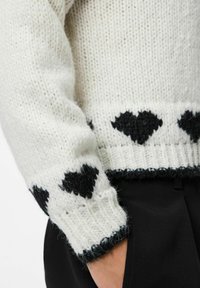 Valge kudumisoa varrukas mustade südame mustritega, kantud mustade pükste juurde, käsi juhuslikult taskusse pandud.