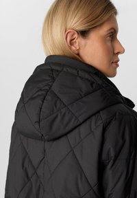 Schwarze gesteppte Jacke mit Kapuze, die ein Rautenmuster und eine matte Textur aufweist, mit sichtbaren Nähdetails und einer taillierten Silhouette.