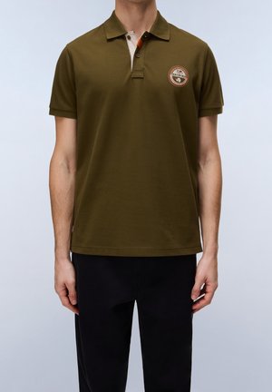 Homme portant un polo manches courtes vert olive avec une patte de boutonnage et un écusson rond sur la poitrine, associé à un pantalon noir.
