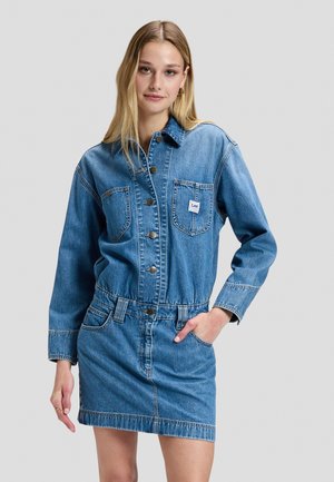Junge Frau trägt ein blaues Jeanskleid mit Fronttaschen und Knöpfen, steht mit einer Hand in der Tasche vor einem einfarbigen Hintergrund.