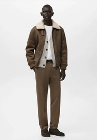 Giacca di shearling marrone con un cardigan a bottoni sotto, maglione crema e pantaloni marroni su misura. Le calzature sono di un marrone scuro.