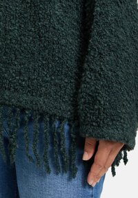 Dunkelgrüner, strukturierter Pullover mit fransigem Saum. Aus dickem Stoff und mit lockerer Passform. Getragen über blauen Jeans. Hand an der Seite resting.