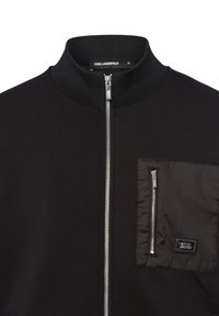 Zwarte jas met een hoge kraag, ritssluiting aan de voorkant en een ritszak. Gemaakt van zacht materiaal met een gladde, strakke afwerking.