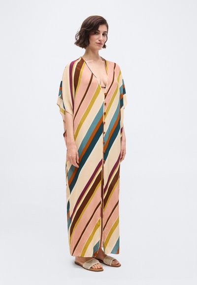 Γυναίκα που στέκεται φορώντας ένα μακρύ, φαρδύ καφτάνι με πολύχρωμες ρίγες και κουμπιά, καθώς και πλεκτά σανδάλια χωρίς λουράκια, σε λευκό φόντο.