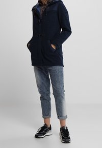 Mörkblå kappa med texturerad yta och mörka axlar, med knappstängning och sidfickor, kombinerad med ljusa, lätt urblekta jeans och svarta sneakers.