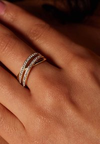 Goldfarbener Ring mit zwei wellenförmigen Bändern. Ein Band hat eine Reihe von kleinen, klaren Edelsteinen, die dem glatten Material und Design Glanz verleihen.