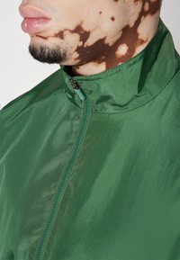 Zelená, lehká bunda s vysokým límcem, s předním zipem a lesklou, reflexní texturou. Detail šití u zipu.