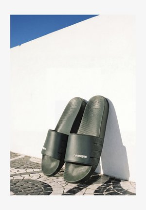 Sandali Havaianas neri a ciabatta appoggiati a un muro bianco su pavimentazione a mosaico nero e bianco sotto un cielo azzurro limpido.