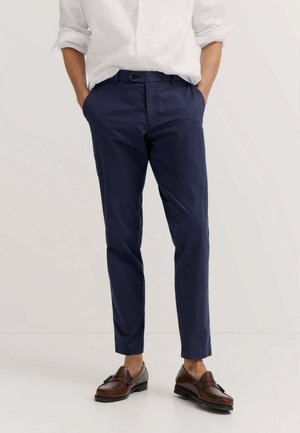 Uomo che indossa pantaloni slim fit blu navy, camicia bianca a maniche lunghe con le maniche rimboccate e mocassini in pelle marrone, mani in tasca.