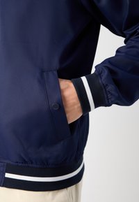 Navy blue lightweight jacket with white striped ribbed cuffs and waistband. Features a side pocket and smooth, glossy finish.