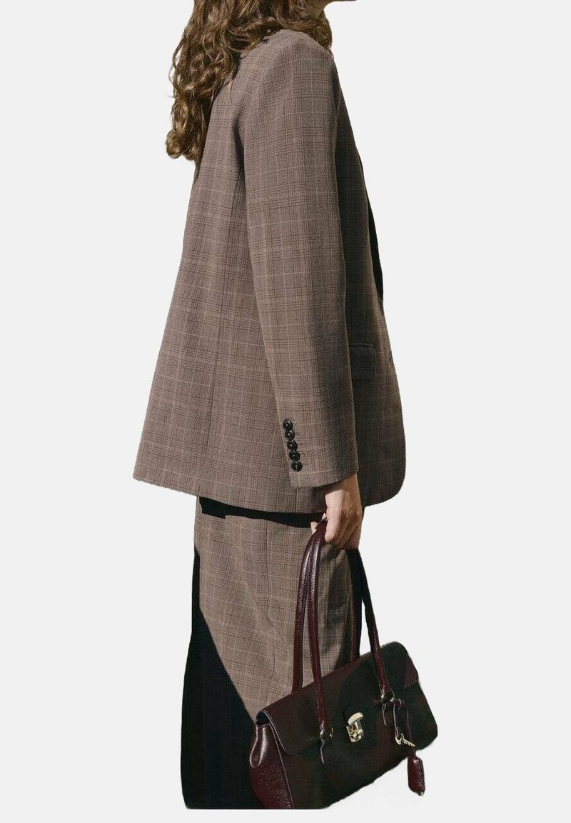 Femme portant un blazer et une jupe à carreaux marron tenant un sac à main en cuir marron foncé sur un fond uni.