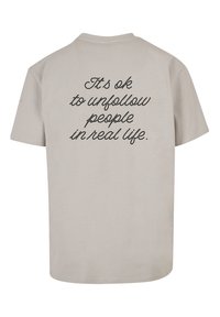 Beige bomulds t-shirt med en afslappet pasform. Bagsiden har sort kursiv tekst, der siger "Det er okay at unfollow folk i det virkelige liv."