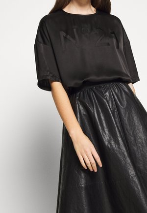 Femme portant un T-shirt ample en satin noir avec "N° 21" embossé et une jupe en cuir noire taille haute sur un fond clair.