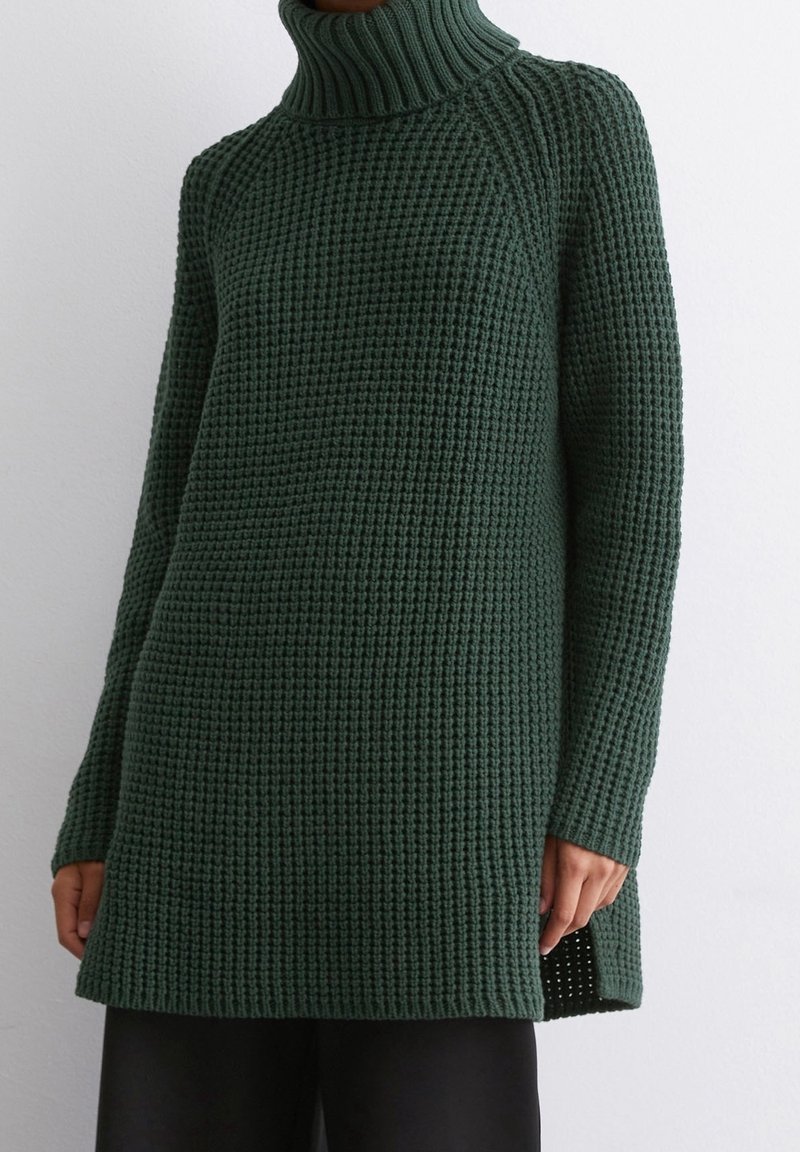 Grüner gestrickter Rollkragenpullover mit strukturiertem Muster, langen Ärmeln und seitlichen Schlitzen. Der Stoff scheint weich und schwer zu sein.