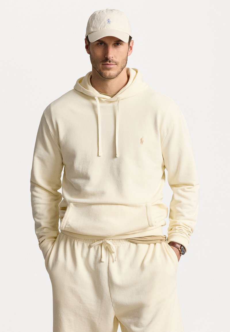 Krämfärgad hoodie och matchande träningsoverallsbyxor i mjukt tyg. Hoodien har en framficka och dragsnören; logotyp på bröstet och på hatten.