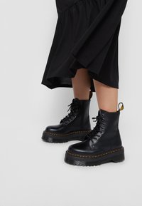 Bottes de combat en cuir noir avec une semelle plateforme épaisse, un bout rond, des lacets ronds et des détails de surpiqûres jaunes. Portées avec un bas de robe noire.