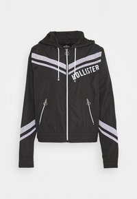 Mustapuku tuulitakki, jossa on valkoiset chevron-raidat, etuvetoketju, sivutaskut ja huppu. Rinnassa on "HOLLISTER"-logo.