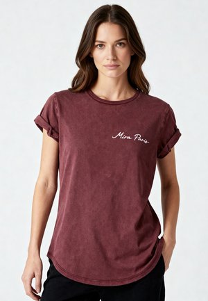 Femme aux cheveux bruns portant un T-shirt bordeaux à manches roulées avec le texte « Mira Paris », posant avec une main dans la poche sur un fond uni.