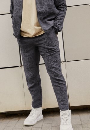 Person iført teksturerede mørkegrå Nike bukser og jakke med beige skjorte og hvide Nike sneakers stående på fortovet.