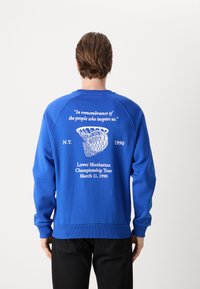 Blauwe sweatshirt met lange mouwen, voorzien van witte tekst en een afbeelding op de achterkant. Zachte stof met geribde manchetten en zoom.