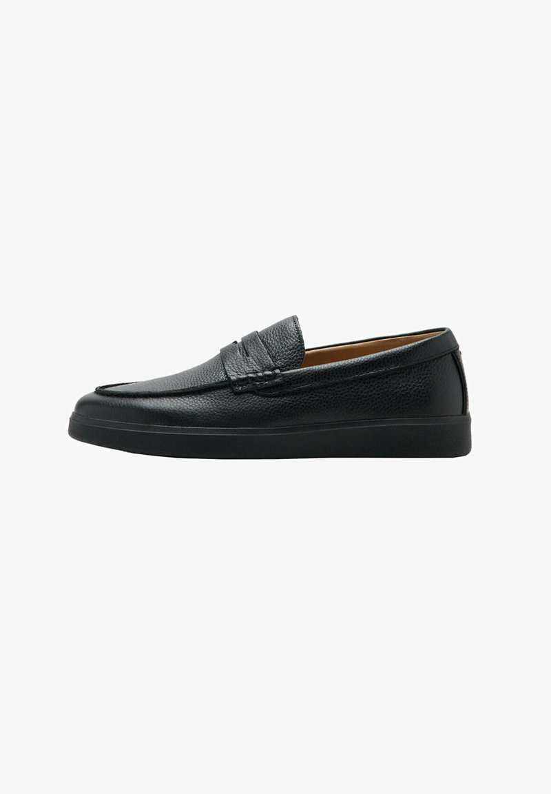 Mocassin en cuir noir sans lacets avec une surface texturée, une semelle plate basse et une lanière décorative sur le dessus.