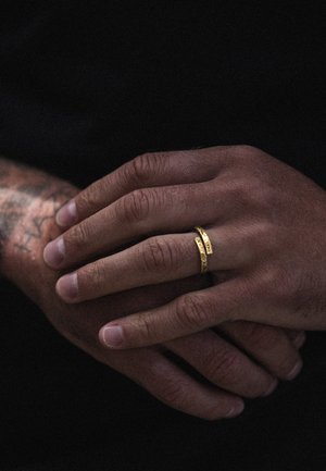 Gold rings with textured surfaces placed on intertwined hands, showcasing a smooth skin texture and detailed wrist tattoos.
