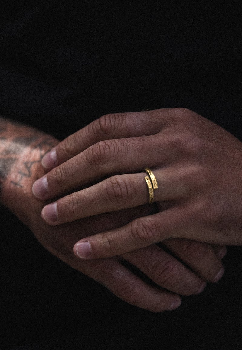 Gold rings with textured surfaces placed on intertwined hands, showcasing a smooth skin texture and detailed wrist tattoos.