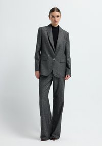 Grauer Anzug mit glitzernder Textur, bestehend aus einem Ein-Knopf-Blazer und weiter Hose. Über einem schwarzen Oberteil getragen, ergänzt durch einen eleganten Stil.
