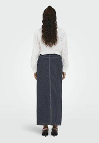 Gonna lunga blu navy con righe bianche verticali, vita alta, silhouette aderente e spacco posteriore. Abbinata a una camicia bianca a maniche lunghe.