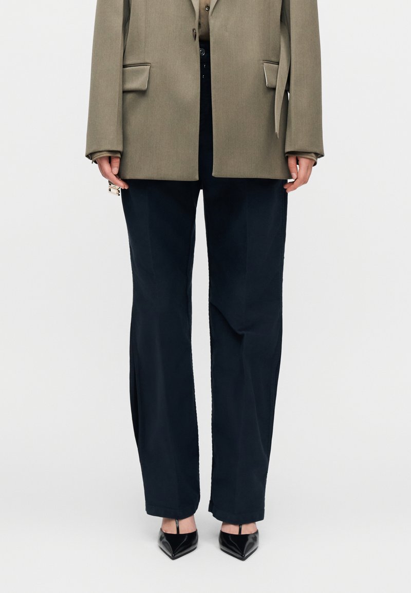 Oseba v olivno zeleni jopiči, črnih širokih hlačah in črnih čevljih s koničastim prstom, stoji pred enobarvnim belim ozadjem.