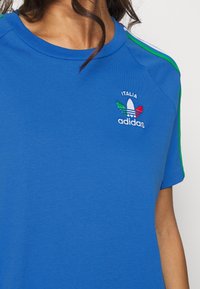 Modré bavlněné tričko s krátkými rukávy, zelenými a bílými pruhy na ramenech a malým vyšívaným logem s nápisy "Italia" a "adidas".