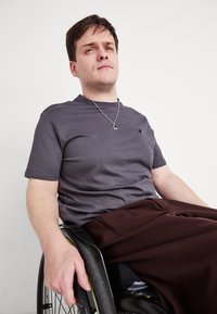 Γκρίζο μπλουζάκι με κοντό μανίκι, με ένα μικρό λογότυπο στο στήθος, συνδυασμένο με καφέ παντελόνι. Ένα χέρι στηρίζεται στο τιμόνι ενός αναπηρικού καροτσιού.