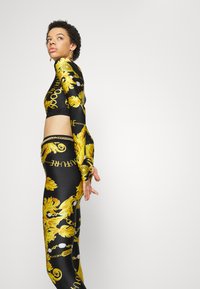 Prilagođena crna majica dugih rukava i odgovarajuće hlače s odvažnim zlatnim cvjetnim i lancem uzorcima, sjajnog izgleda i izrezom na trbuhu.