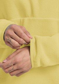 Tessuto di felpa giallo con una texture liscia, che mostra un polsino in fase di regolazione. Dettagli notevoli includono le cuciture e i tatuaggi sulle mani.