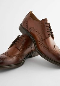Chaussures en cuir marron avec des détails brogue, bout rond et semelle en caoutchouc noire. Présente un système de laçage et des accents gaufrés.