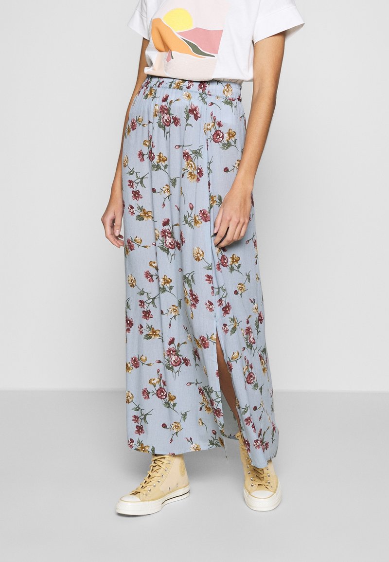 Femme portant un t-shirt blanc et une jupe longue fleurie bleu clair avec une fente latérale, associée à des baskets montantes beiges.