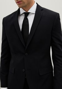 Completo nero con una vestibilità su misura, design a un bottone, reverse a falda, due bottoni e tasche laterali. Abbinato a una camicia bianca e una cravatta nera.