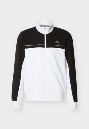 Λευκό και μαύρο μπουφάν με φερμουάρ και ψηλό γιακά, μαύρο πάνω μέρος στο στήθος και τα μανίκια, λευκό στο κάτω μέρος του σώματος, με το λογότυπο του κροκοδείλου της Lacoste στο στήθος.