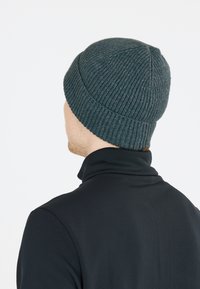 Grå stickad mössa med ribbat mönster, konisk form och rundad topp, som bärs av en person i ett plagg med hög krage i svart.