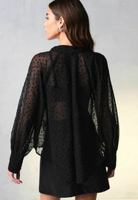 Sort chiffonbluse med hvide polka prikkers, lange, blafrende ærmer og knapmanchetter; falder løst med en afrundet kant, parret med en lædernederdel.