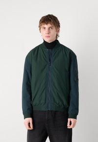 Jachetă bomber verde cu mâneci contrastante de navy, material ușor, închidere cu fermoar, guler ribbed și buzunar pe mâneca stângă.