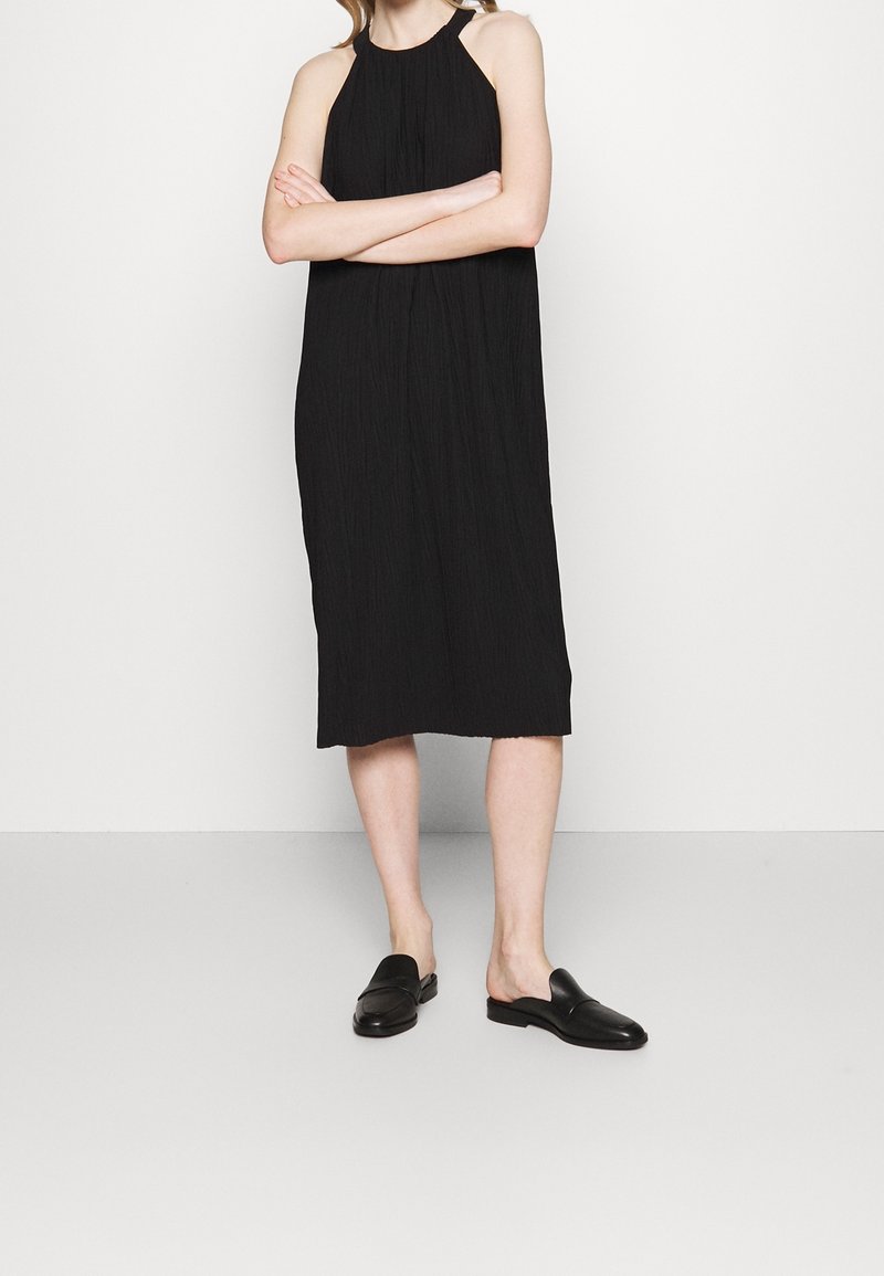 Musta halterneck-mekko, jossa on rento istuvuus, teksturoitu kangas ja polvipituinen helma. Yhdistetty mustiin liukuhihnakenkiin.