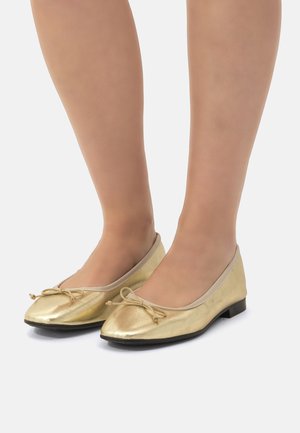 Bailarinas doradas con un acabado metálico suave, punta redondeada y un pequeño lazo decorativo en la parte delantera. Presentan un bajo tacón negro.