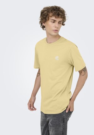 Joven con cabello rizado que lleva una camiseta amarillo pálido y vaqueros grises, mirando hacia su izquierda con una expresión neutral.